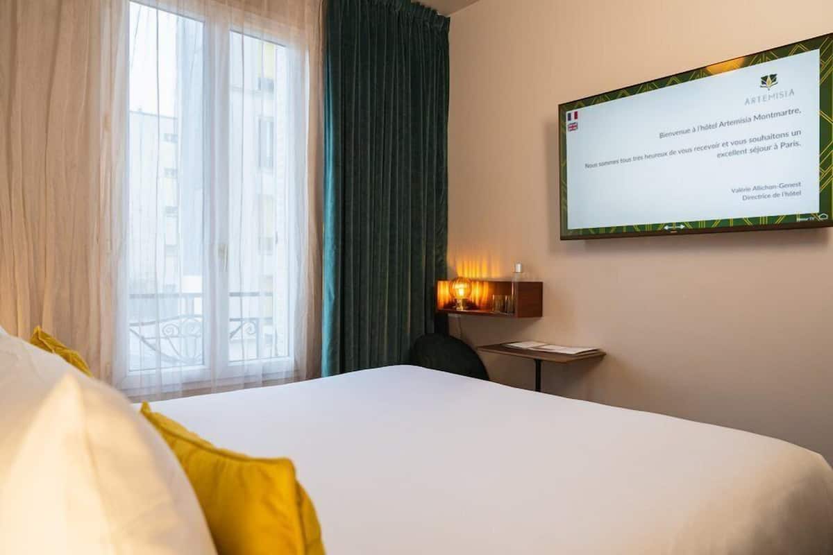 Una camera d'albergo con un letto, una scrivania, una sedia, una finestra con tende e uno schermo a parete che mostra un messaggio di benvenuto.