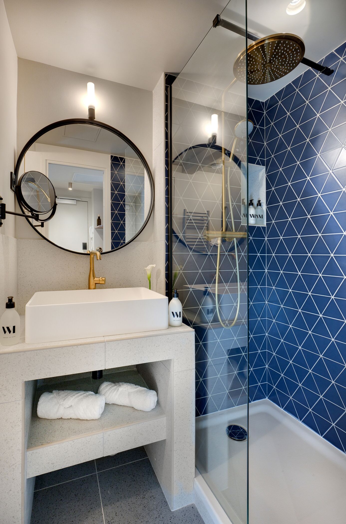 Un baño con lavamanos blanco, grifo dorado y espejo redondo. Cuenta con una ducha con mampara de vidrio, cabezal de ducha manual y un toallero de pared.