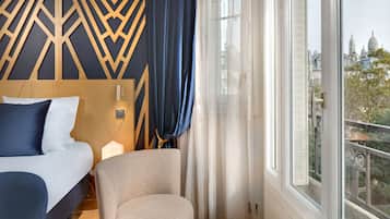 Ein Hotelzimmer mit Bett, Sessel, einem kleinen Tisch mit Vase und einem großen Fenster mit Blick auf Gebäude.