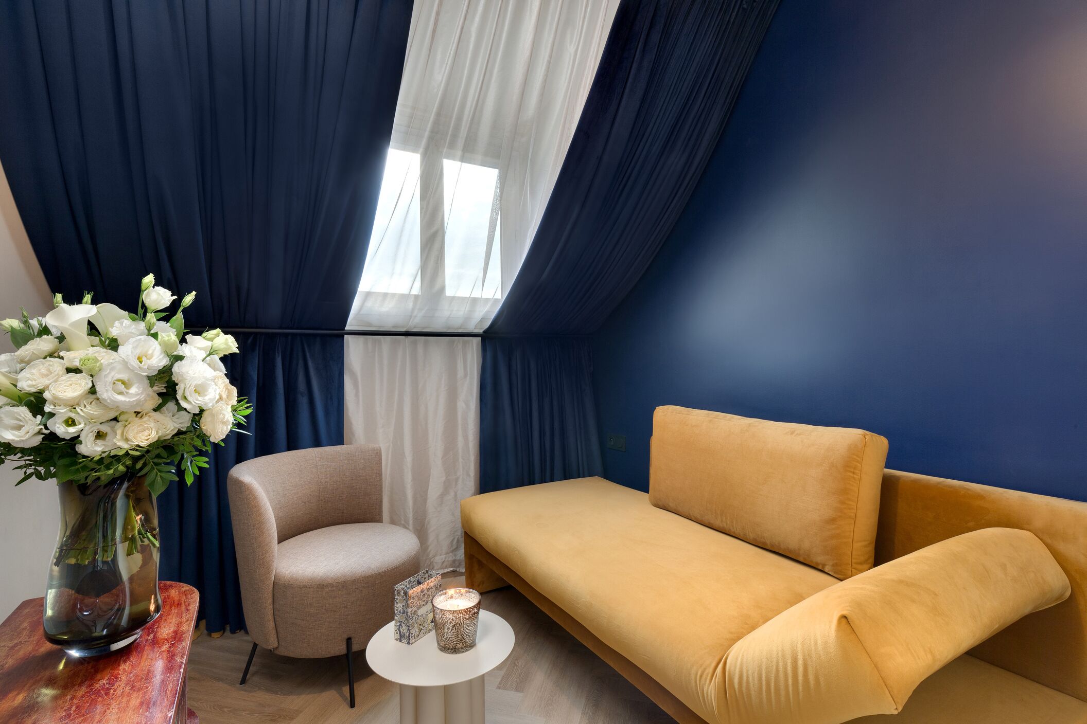 Une pièce avec un mur bleu, un canapé beige, un fauteuil blanc, une table ronde ornée d’un vase de fleurs blanches et une petite bougie.
