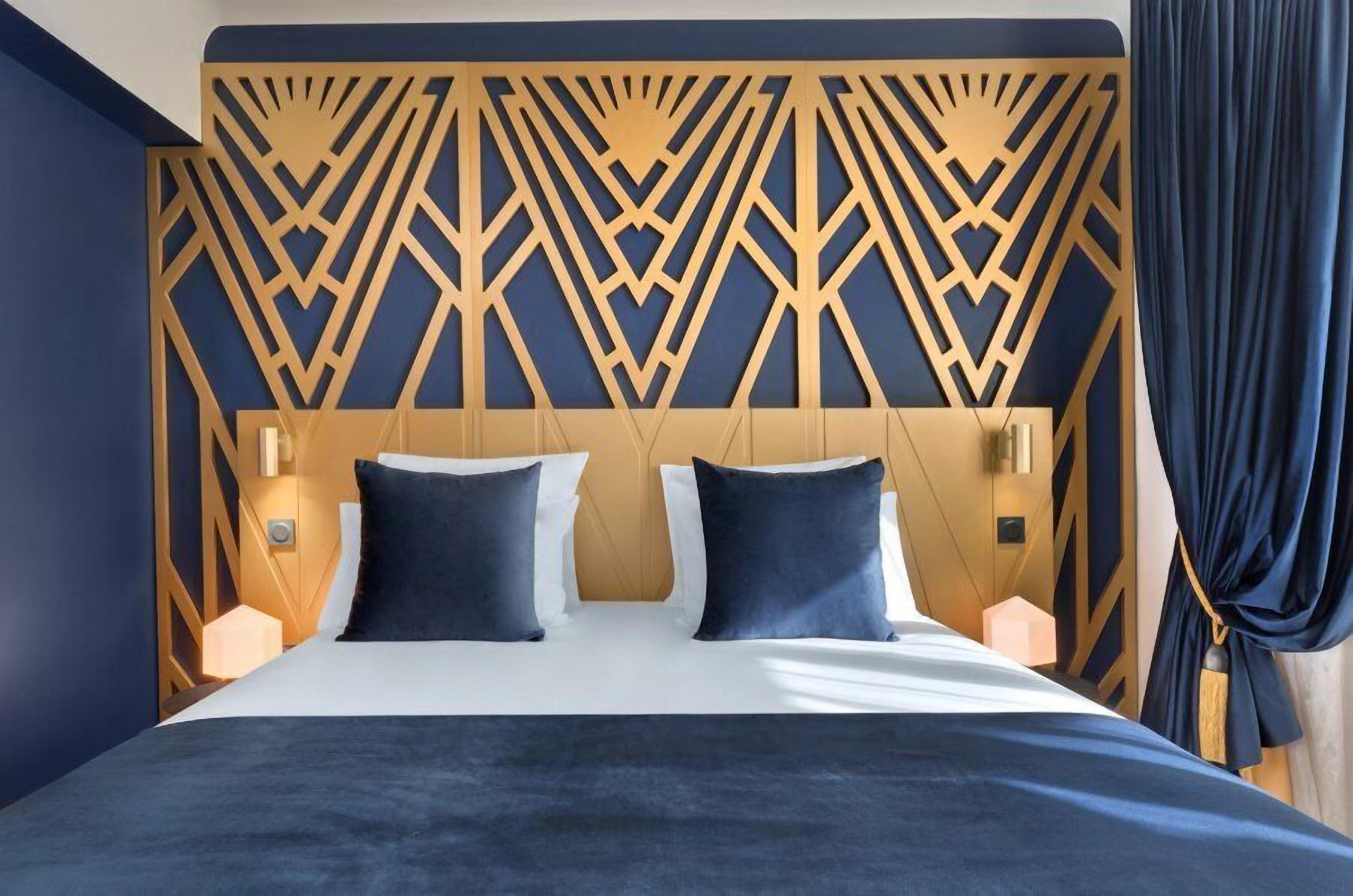 A hotel room with a geometric patterned headboard, a bed with dark bedding, and two bedside lamps.