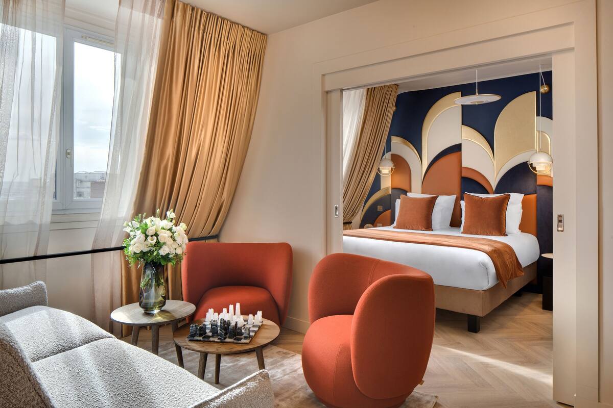 Una camera d'albergo con un letto, due poltrone arancioni, un tavolino rotondo con una scacchiera e un vaso di fiori.