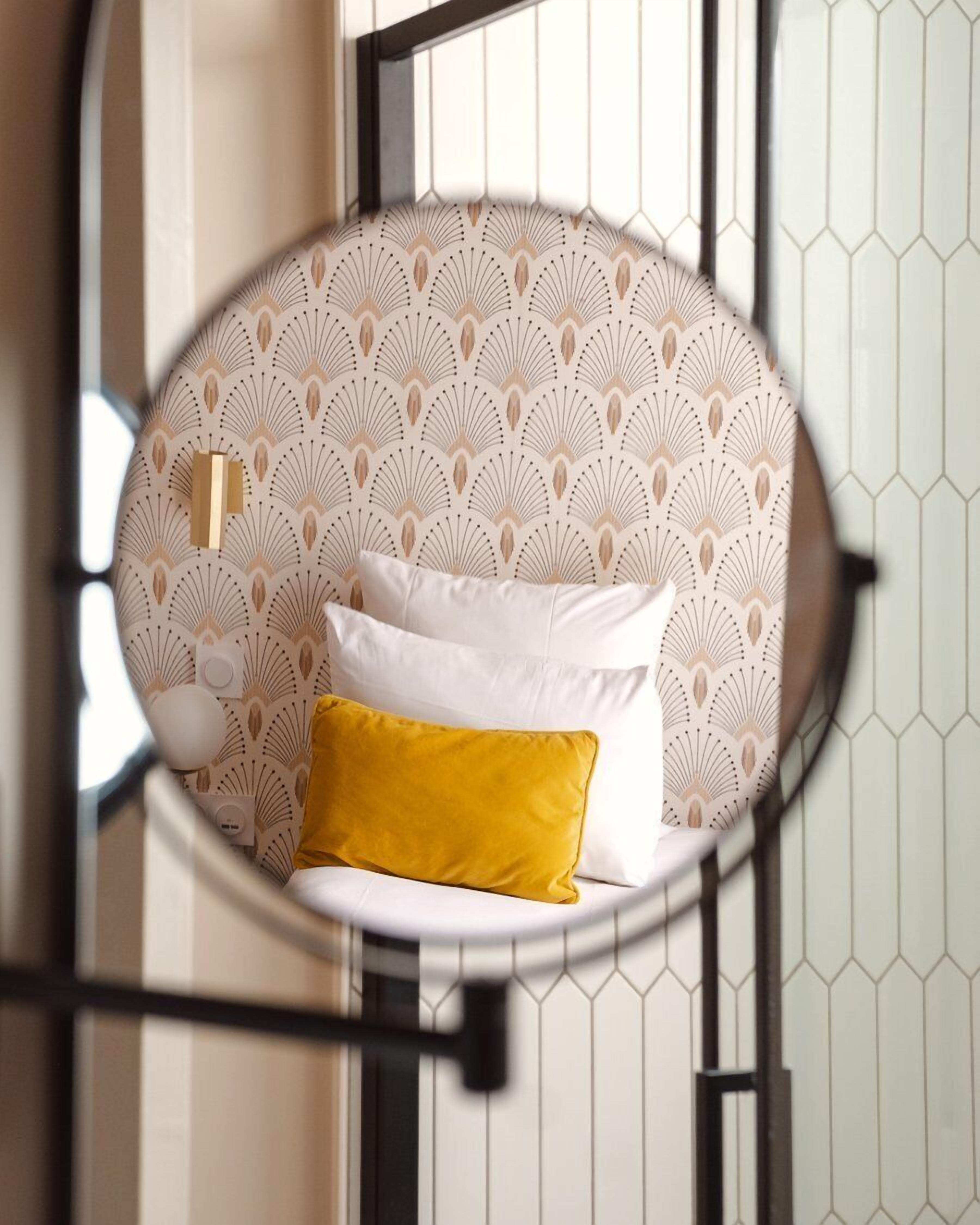 A circular mirror reflecting a bed with white and yellow pillows, a patterned wallpaper, and a hexagonal tile wall.