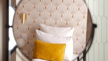 A circular mirror reflecting a bed with white and yellow pillows, a patterned wallpaper, and a hexagonal tile wall.