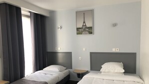 Habitación de hotel con dos camas, una ventana con cortinas, un cuadro en la pared de la Torre Eiffel y una mesita.