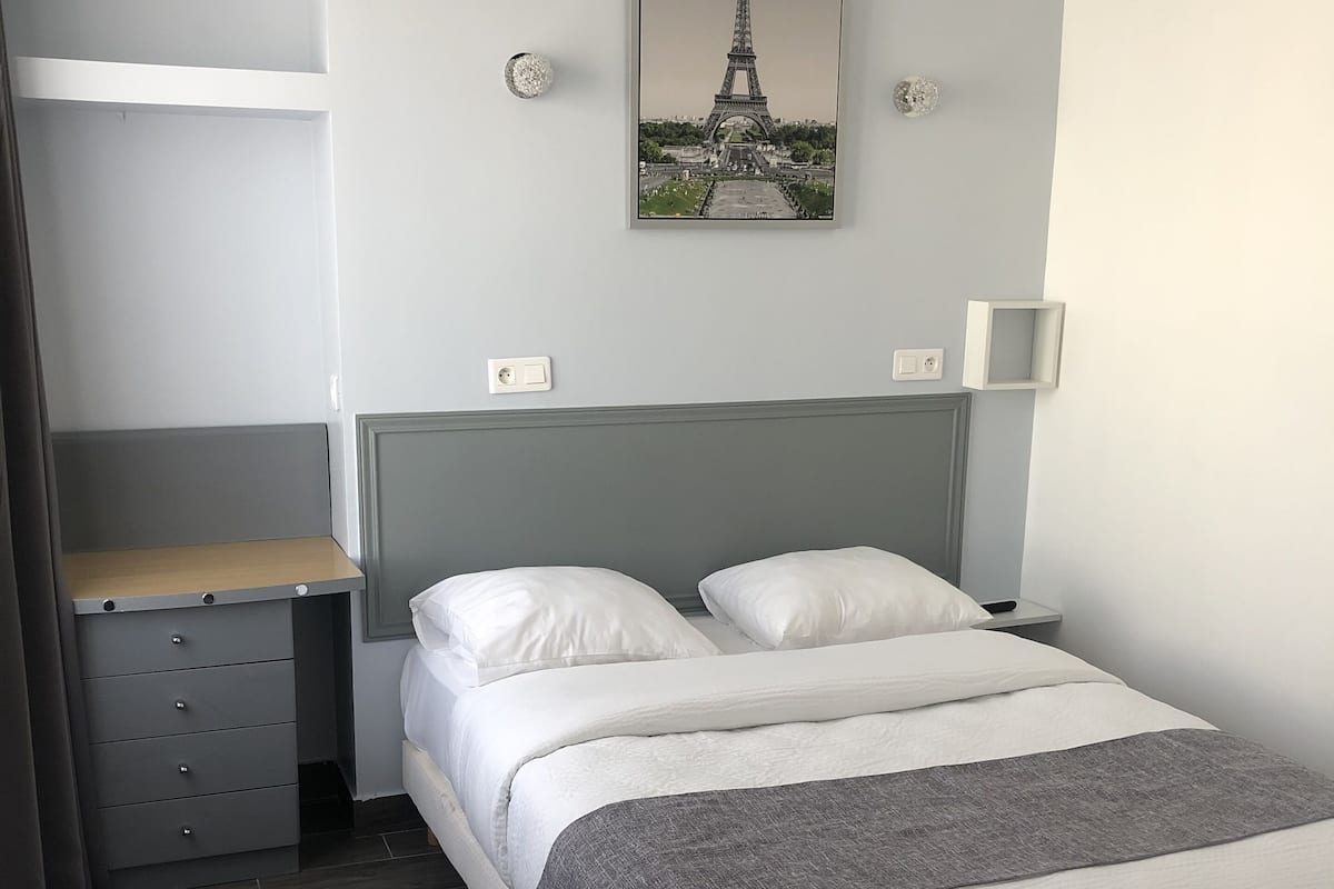 A neatly arranged hotel room with a bed, a desk with drawers, a lamp, and a picture of the Eiffel Tower on the wall.