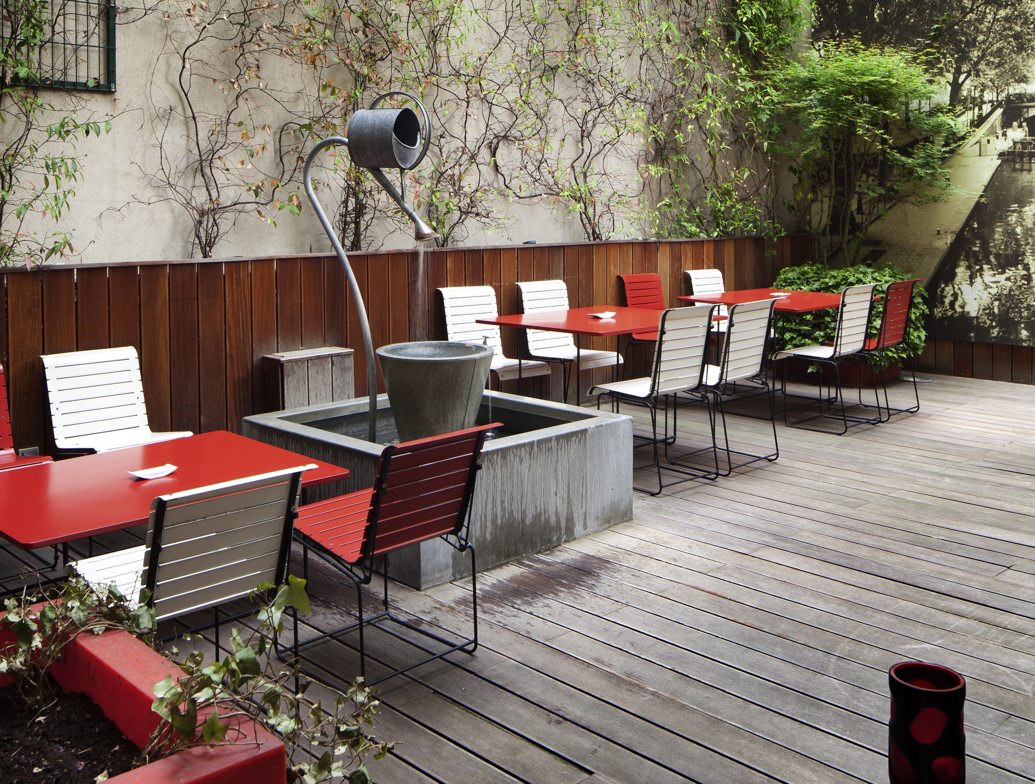 An outdoor patio with red and white tables and chairs, a large planter, and a unique watering can sculpture.