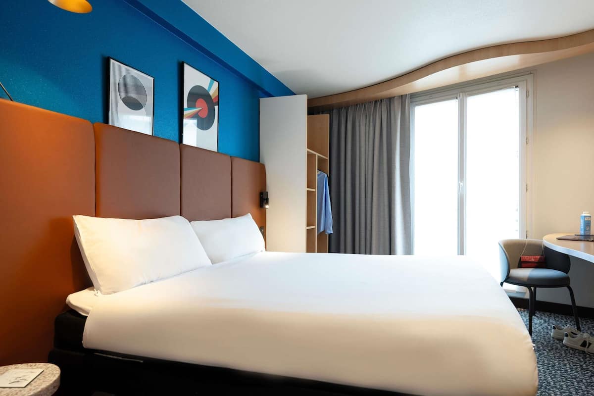 Une chambre d’hôtel avec un grand lit, un bureau et une chaise. Il y a une fenêtre avec des rideaux, un mur décoré d’œuvres d’art et un plafond avec un luminaire.
