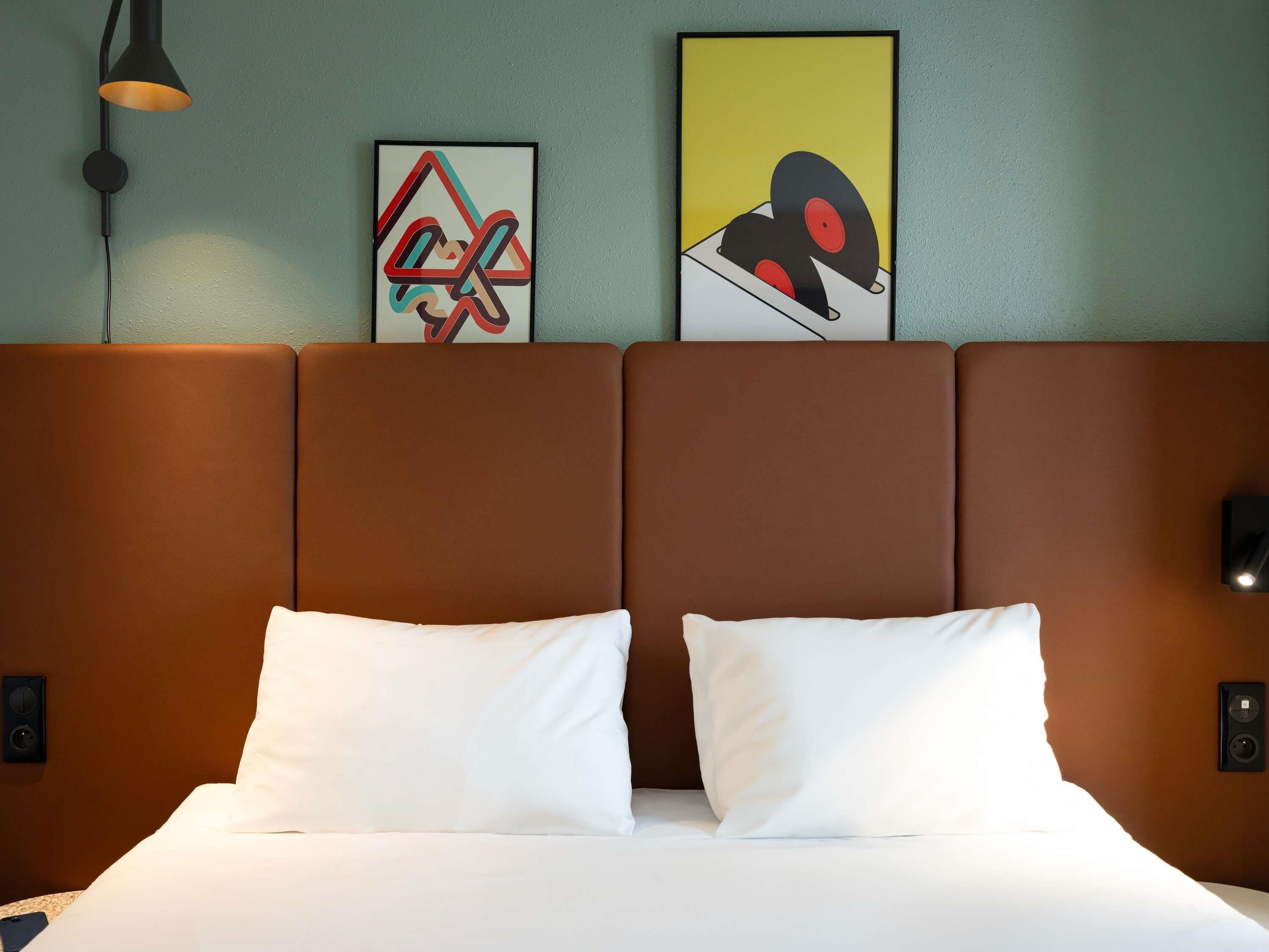 A hotel room with a brown headboard, white pillows, a framed abstract art piece, and a wall-mounted light fixture.