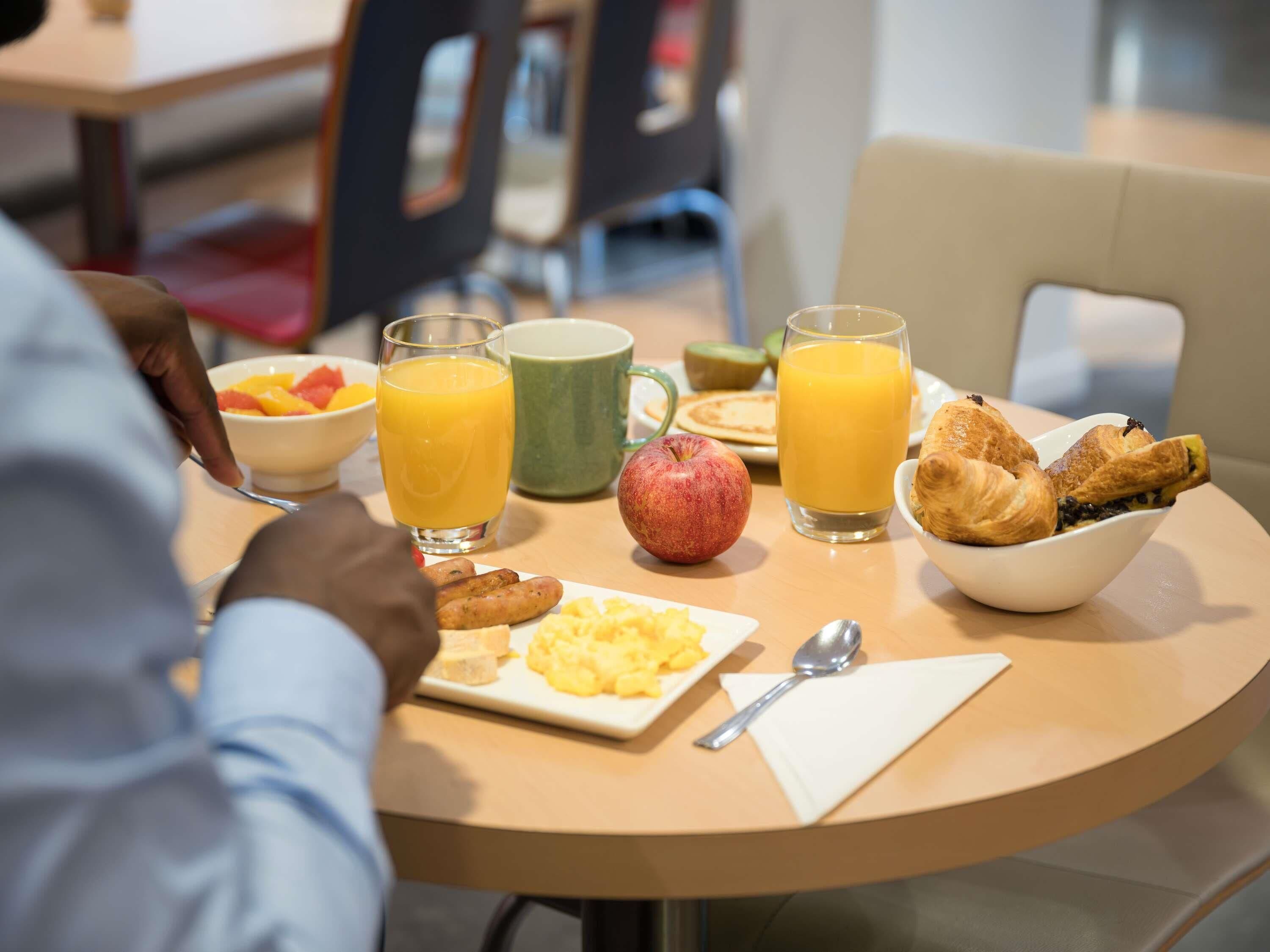 A breakfast table with a plate of scrambled eggs, a bowl of croissants, a glass of orange juice, a green mug, and a bowl of fruit.