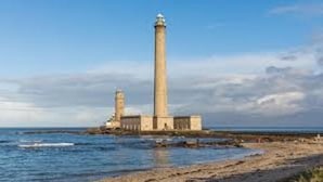 A lighthouse with a tall, slender tower and a shorter structure nearby, situated on a rocky shoreline with the sea in the background.