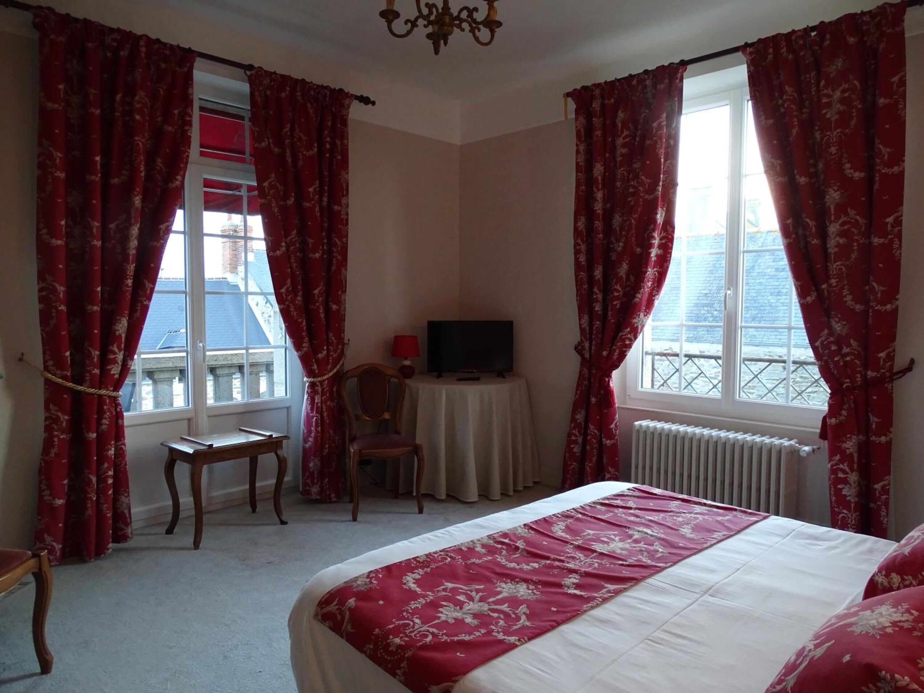 A hotel room with a bed, two chairs, a television on a small table, and large windows with red curtains.