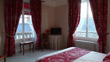 A hotel room with a bed, two chairs, a television on a small table, and large windows with red curtains.