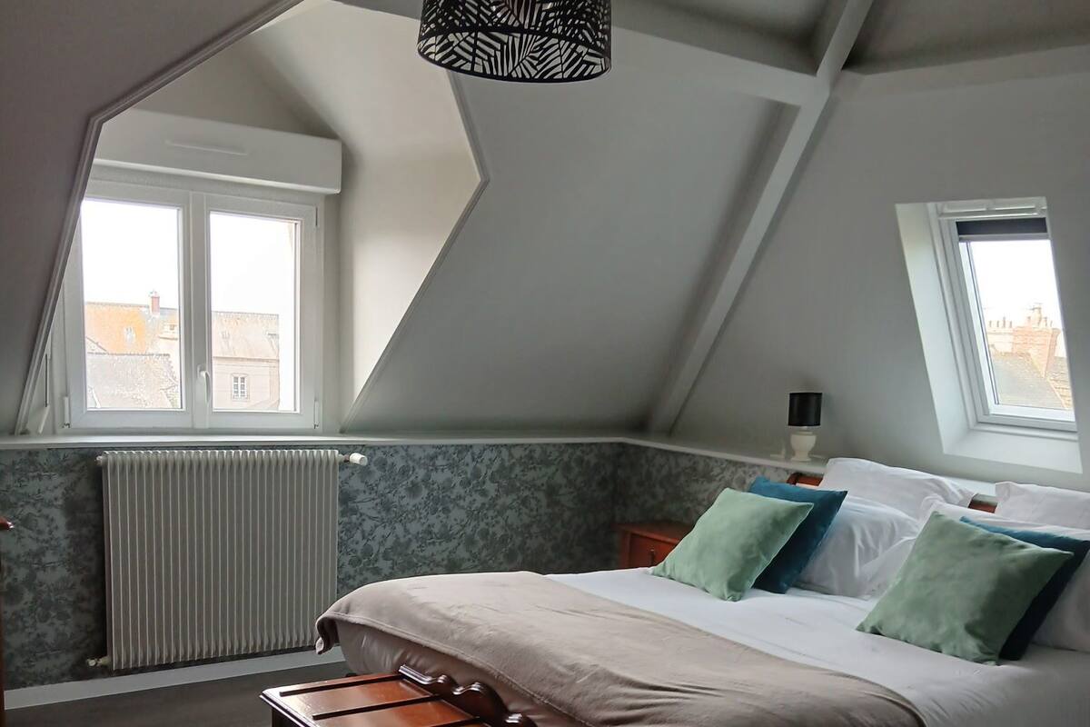 Een slaapkamer met een schuine kap, een bed met kussens, een bankje, een radiator en een raam.