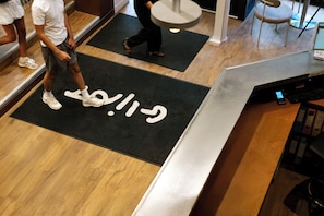 Uma área de recepção com tapete preto com texto branco, piso de madeira e balcão de metal.