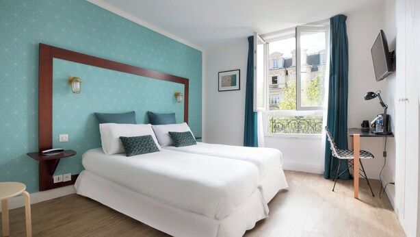 A hotel room with two beds, a desk, a chair, a TV, and a window with a view of buildings.