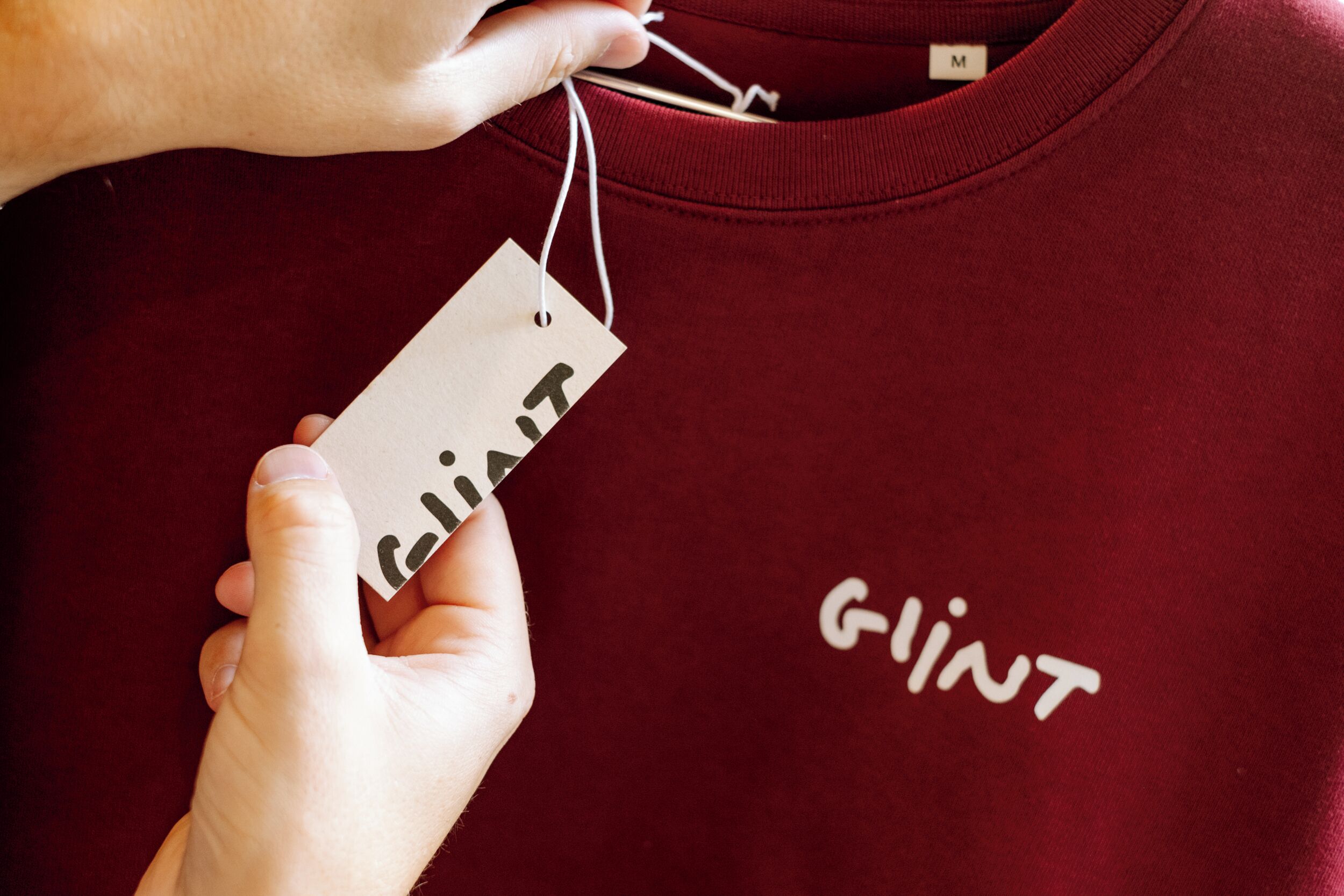 Une main tenant une étiquette sur laquelle est écrit le mot « glint », sur un t-shirt bordeaux où le mot « glint » est également imprimé.