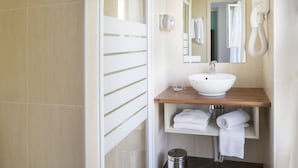 A modern bathroom with a white sink, wooden countertop, and a mirror. There are two white towels on the shelf and a trash bin on the floor.