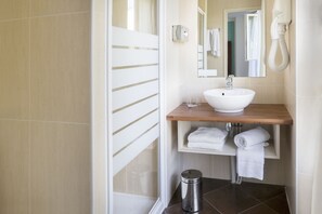 Ein modernes Badezimmer mit einem weißen Waschbecken, einer Holzauskleidung und einem Spiegel. Auf dem Regal liegen zwei weiße Handtücher und ein Mülleimer steht auf dem Boden.