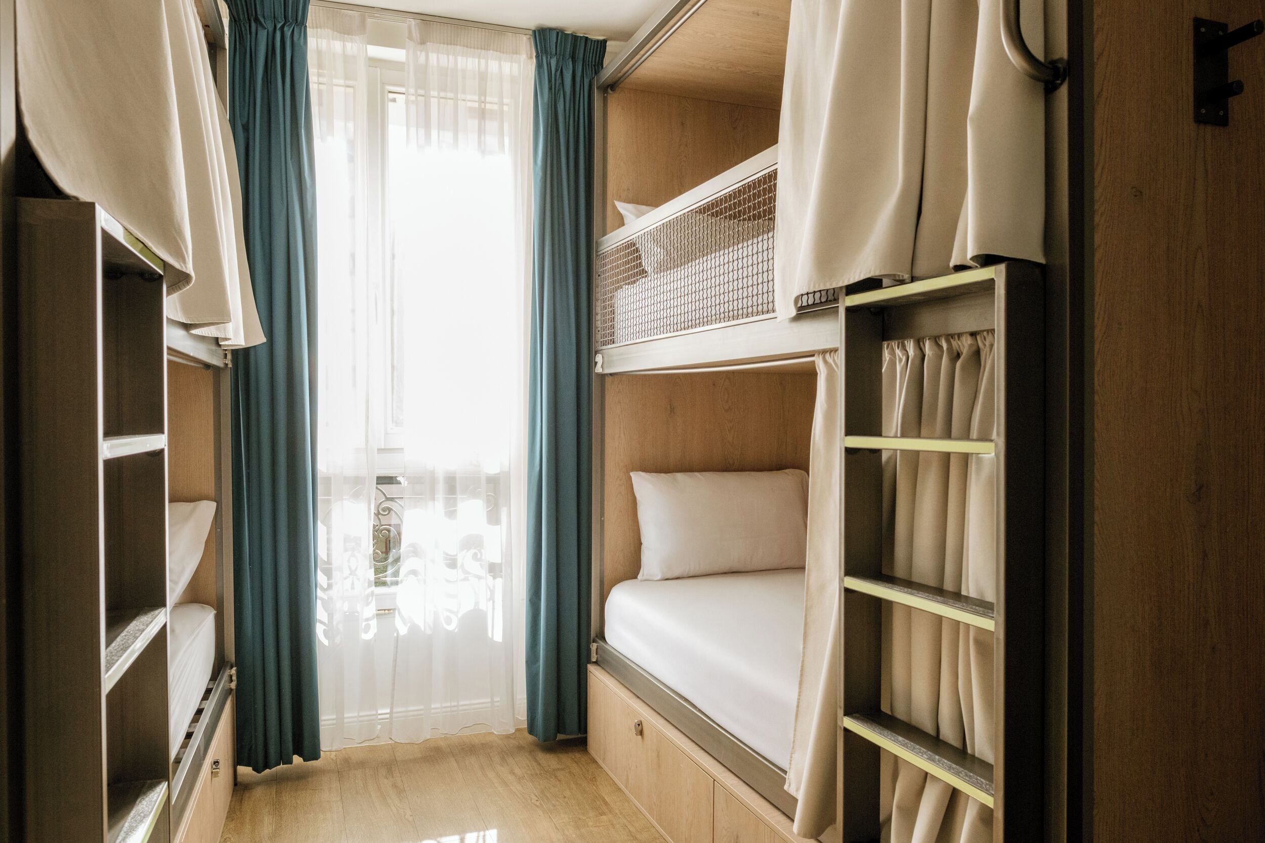 A bunk bed room with a window, curtains, and a wooden floor.