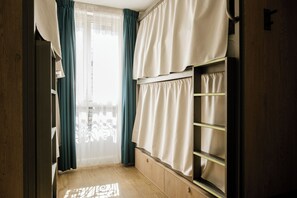 Una habitación con piso de madera, una ventana con cortinas traslúcidas y una estantería empotrada con cortina.