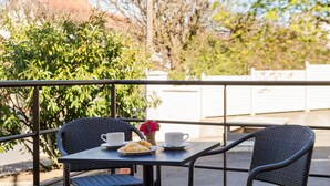 Un balcon avec des fauteuils en osier, une table avec une tasse, une soucoupe et une assiette de nourriture, et une plante en pot.