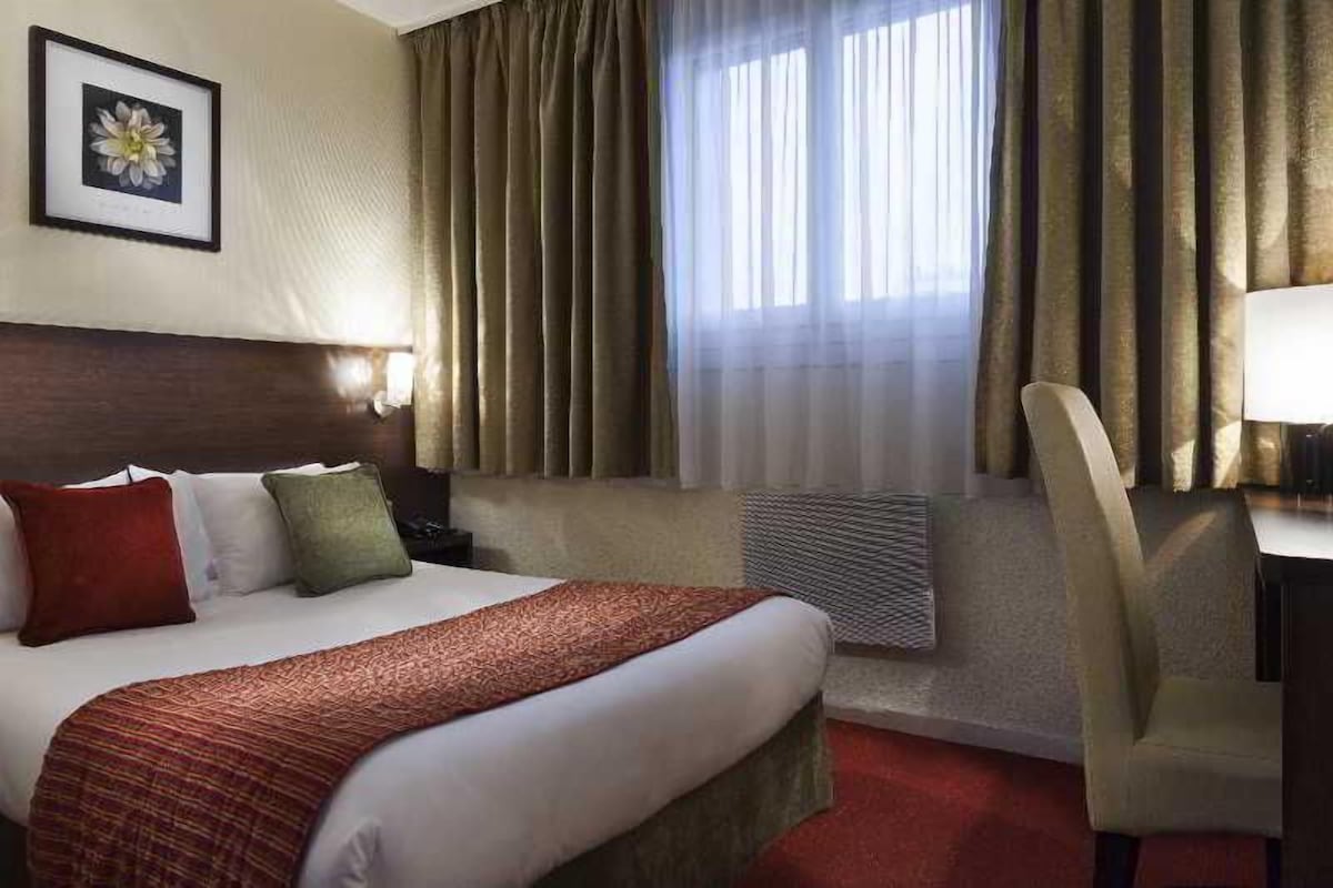 Hotelkamer met een groot bed, een bureau, een stoel, een inlijsting foto aan de muur en een raam met gordijnen.