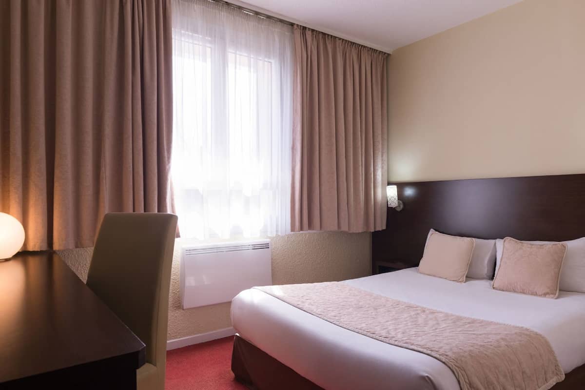 Habitación de hotel con cama, escritorio, silla, ventana con cortinas y lámpara en la pared.