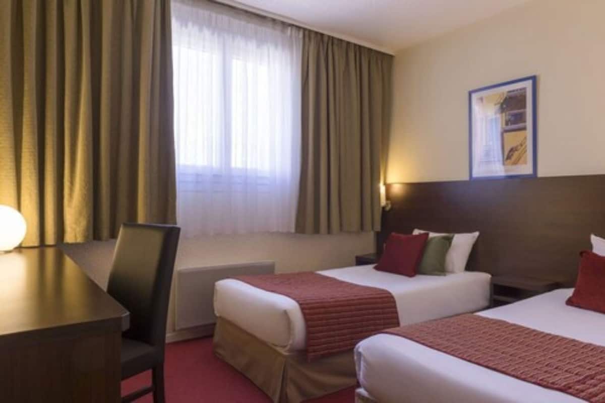 Una habitación de hotel con dos camas, un escritorio, una silla y una ventana con cortinas.