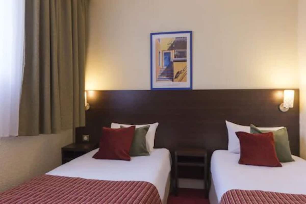 Habitación de hotel con dos camas, cada una con almohada roja y verde, un cuadro enmarcado en la pared y una mesita de noche con lámpara.