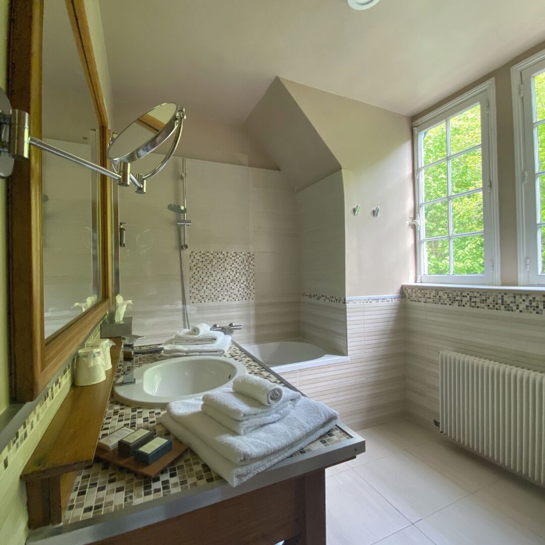 A bathroom with a large mirror, two sinks, a bathtub, and a window with a view of greenery.
