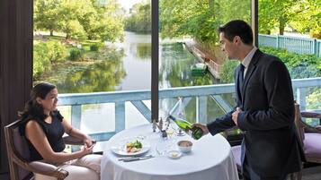 Una mesa de comedor con vistas a un río y un puente, un camarero sirviendo una botella de vino y una mujer sentada a la mesa.