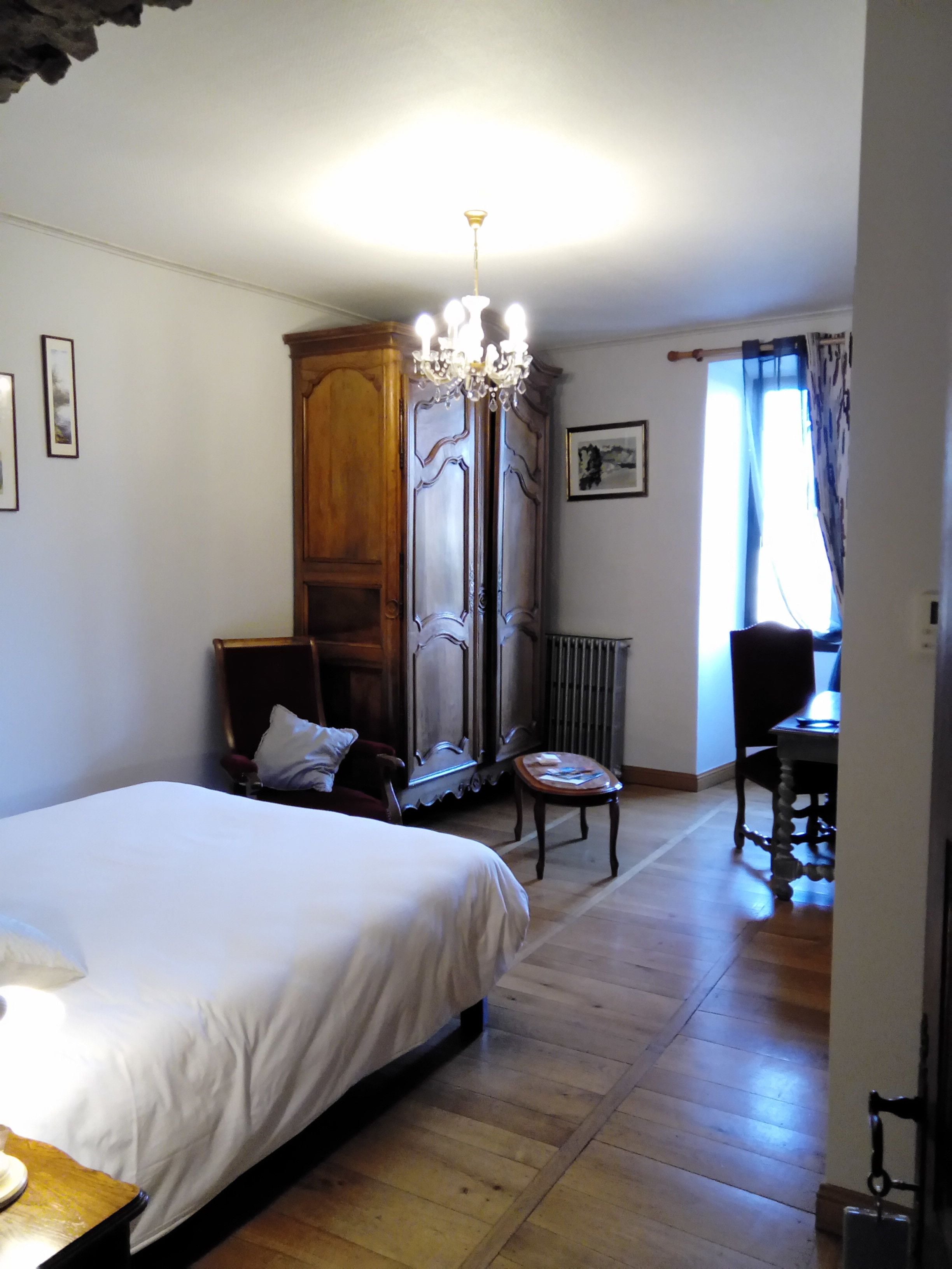 A bedroom with a large bed, a wooden wardrobe, a chair, a table, and a chandelier.