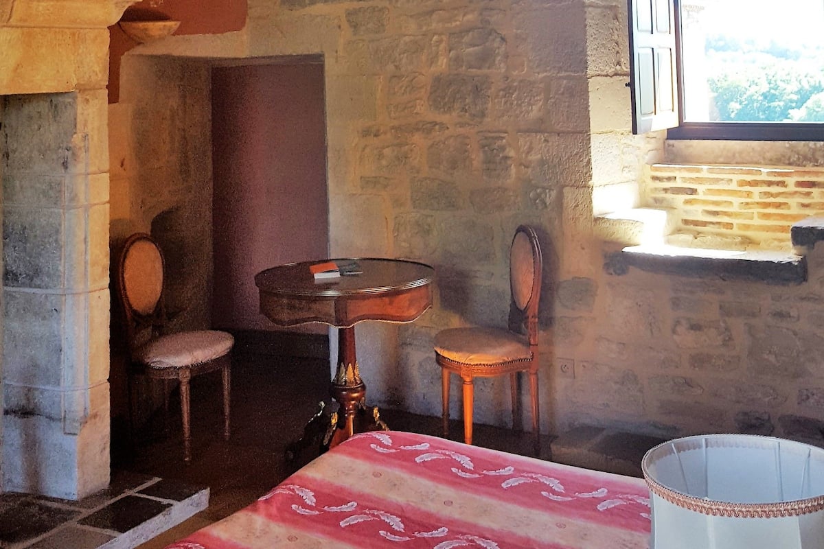 A rustic room with exposed wooden beams, a stone wall, a chandelier, a bed with a patterned bedspread, a table with chairs, and a window with a view.