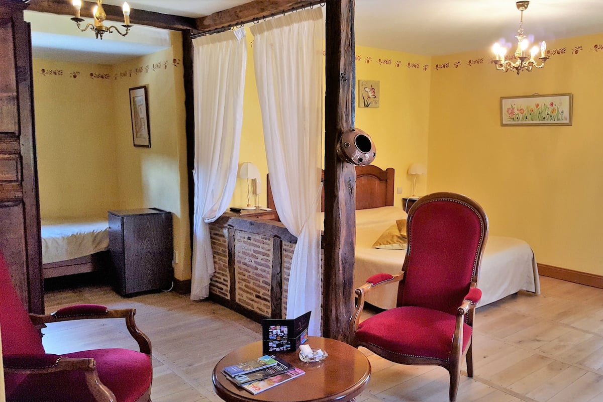 A room with a bed, a wooden chair, a table with a magazine, and a chandelier.