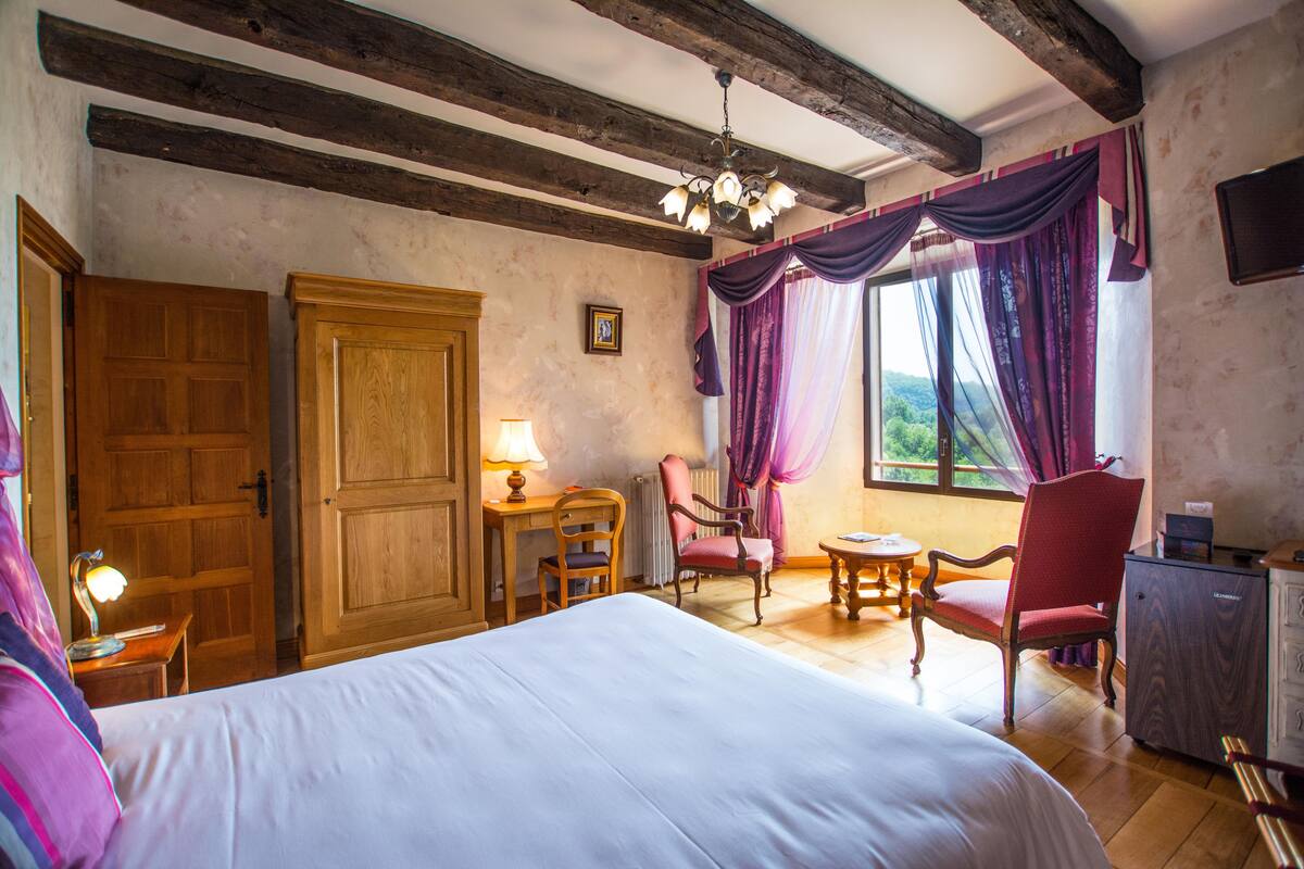 A bedroom with a large bed, wooden furniture, a desk with a lamp, and a view of the outdoors through a window with curtains.