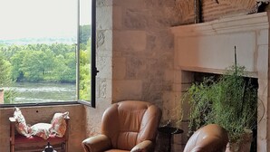 Quarto rústico com lareira, poltronas de couro, uma mesa com bule e vista para um rio e floresta.