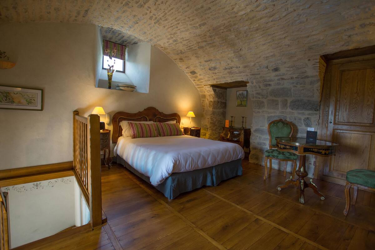 A bedroom with a stone ceiling, a wooden bed, a nightstand, a chair, and a small table.