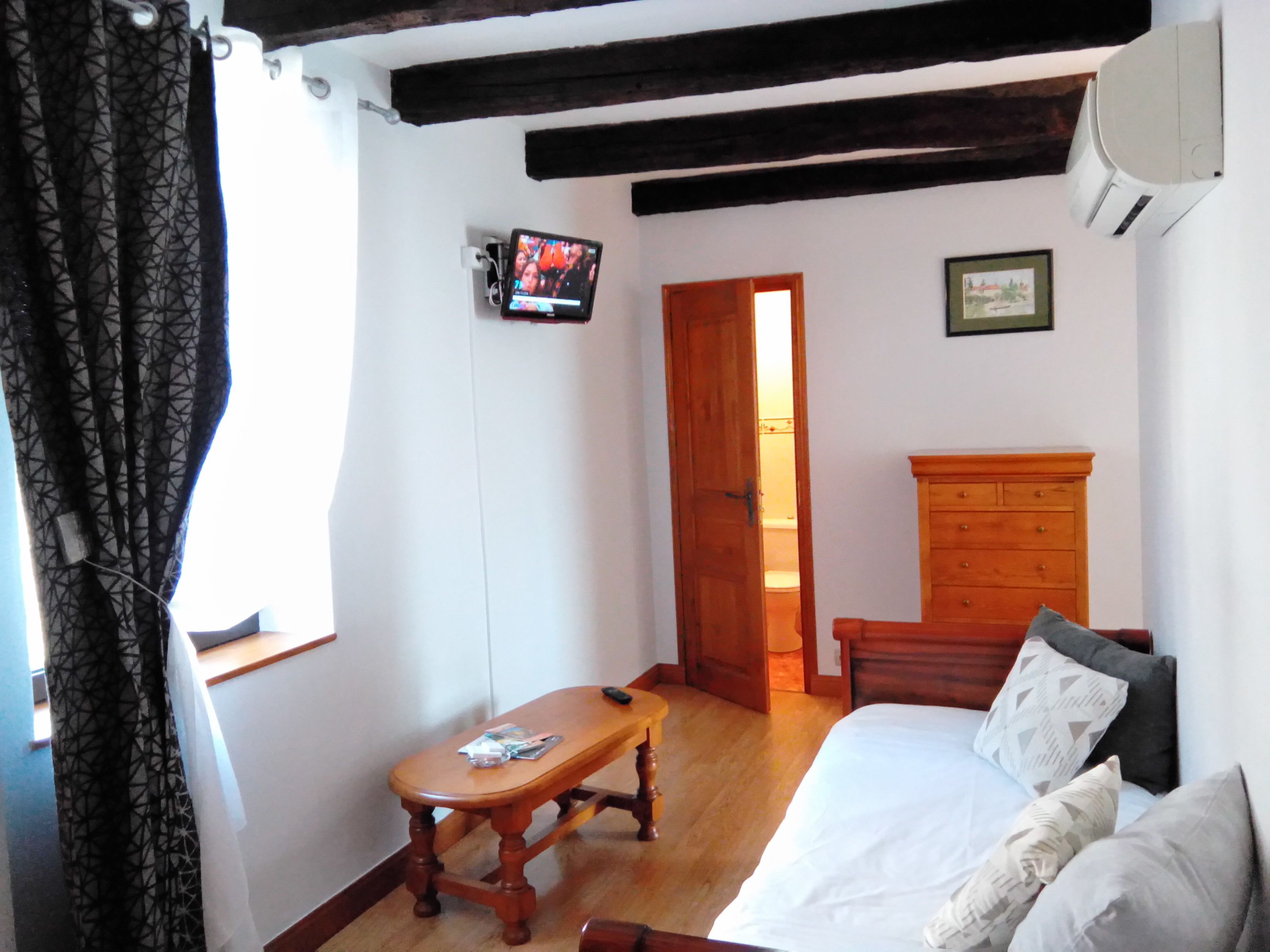A bedroom with a wooden bed, a wooden nightstand, a wooden bench, a television on the wall, and a door leading to another room.