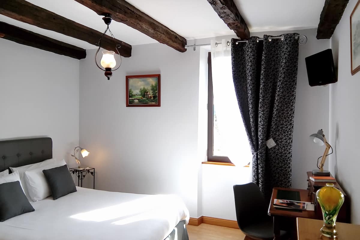 A bedroom with a bed, a desk, a chair, a lamp, a painting, and a window with curtains.