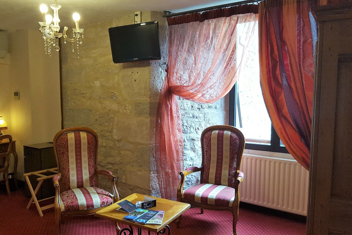 A room with two upholstered chairs, a wooden table with magazines, a television on the wall, and a window with curtains.