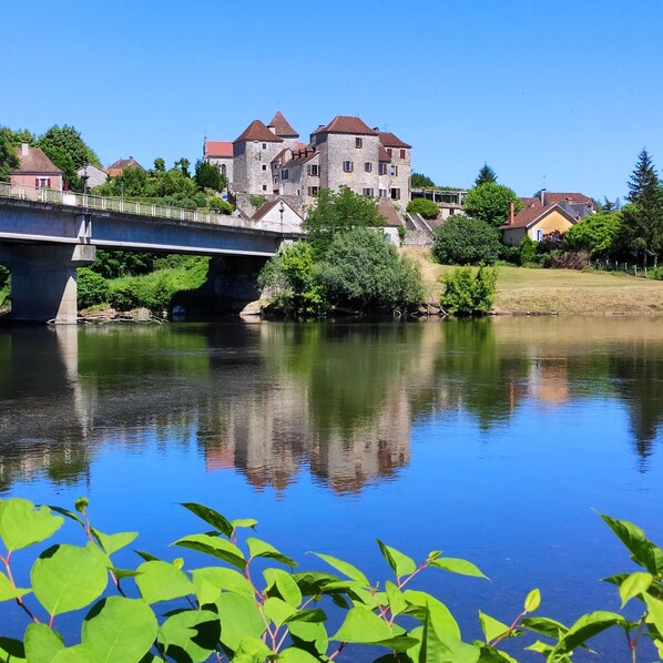 A stone bridge over a calm river, with a castle-like building in the background and lush greenery in the foreground.