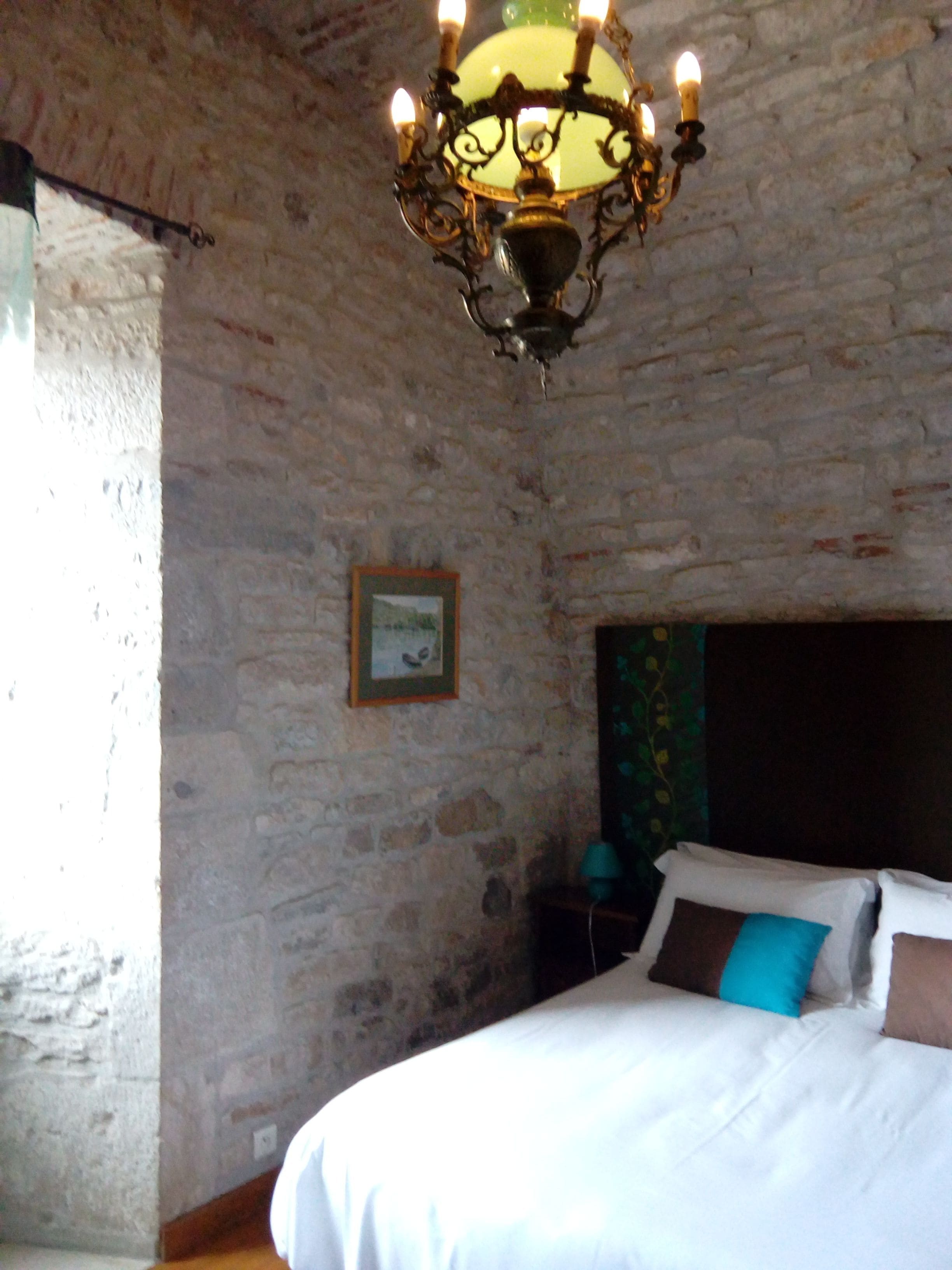 Ein Schlafzimmer mit Steinwand, einer Chandelüster, einem Bett mit weißer und brauner Bettwäsche sowie einem gerahmten Bild an der Wand.