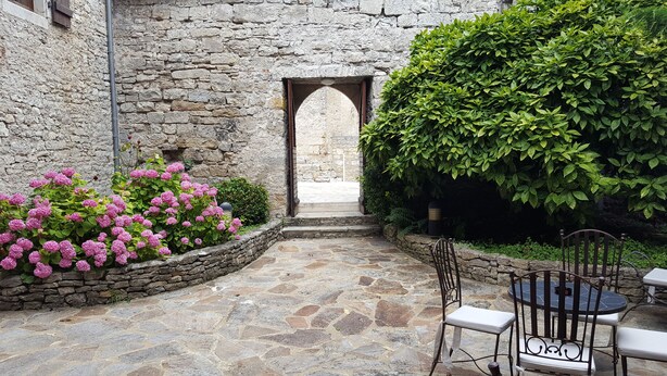 Um pátio de pedra com um arco rústico, um muro de pedra com arbustos de hortênsia rosa e uma mesa de metal com cadeiras.