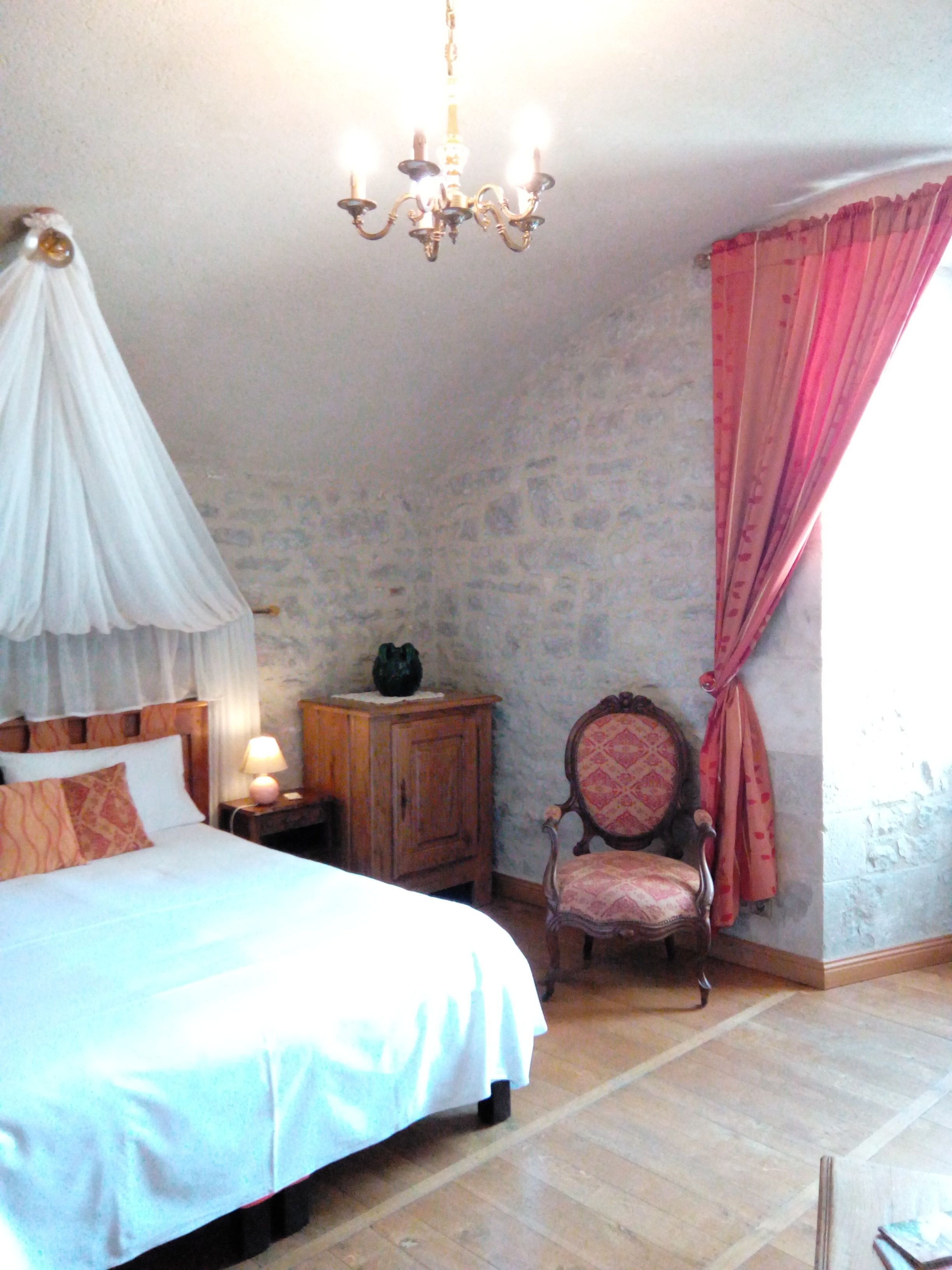 A bedroom with a canopy bed, a wooden nightstand with a lamp, a chair, and a window with pink curtains.
