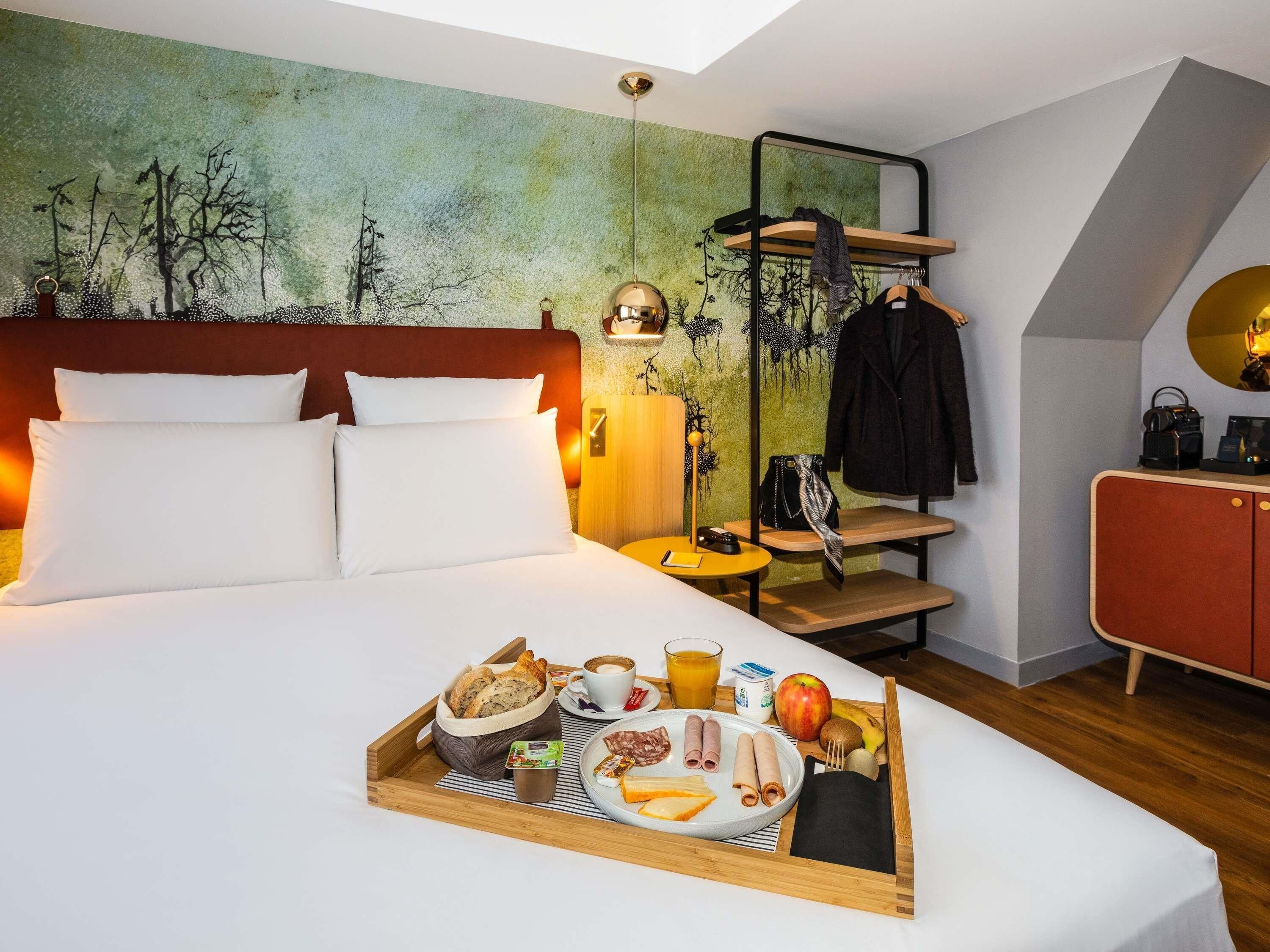 Habitación de hotel con cama, bandeja de desayuno, armario y un mural de un árbol en la pared.