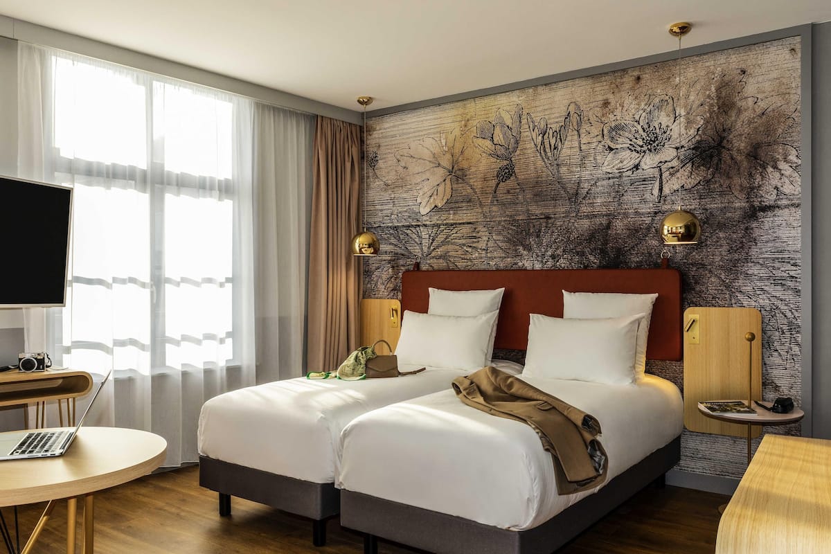 A hotel room with a double bed, a desk with a laptop, a television, and a large window with curtains.