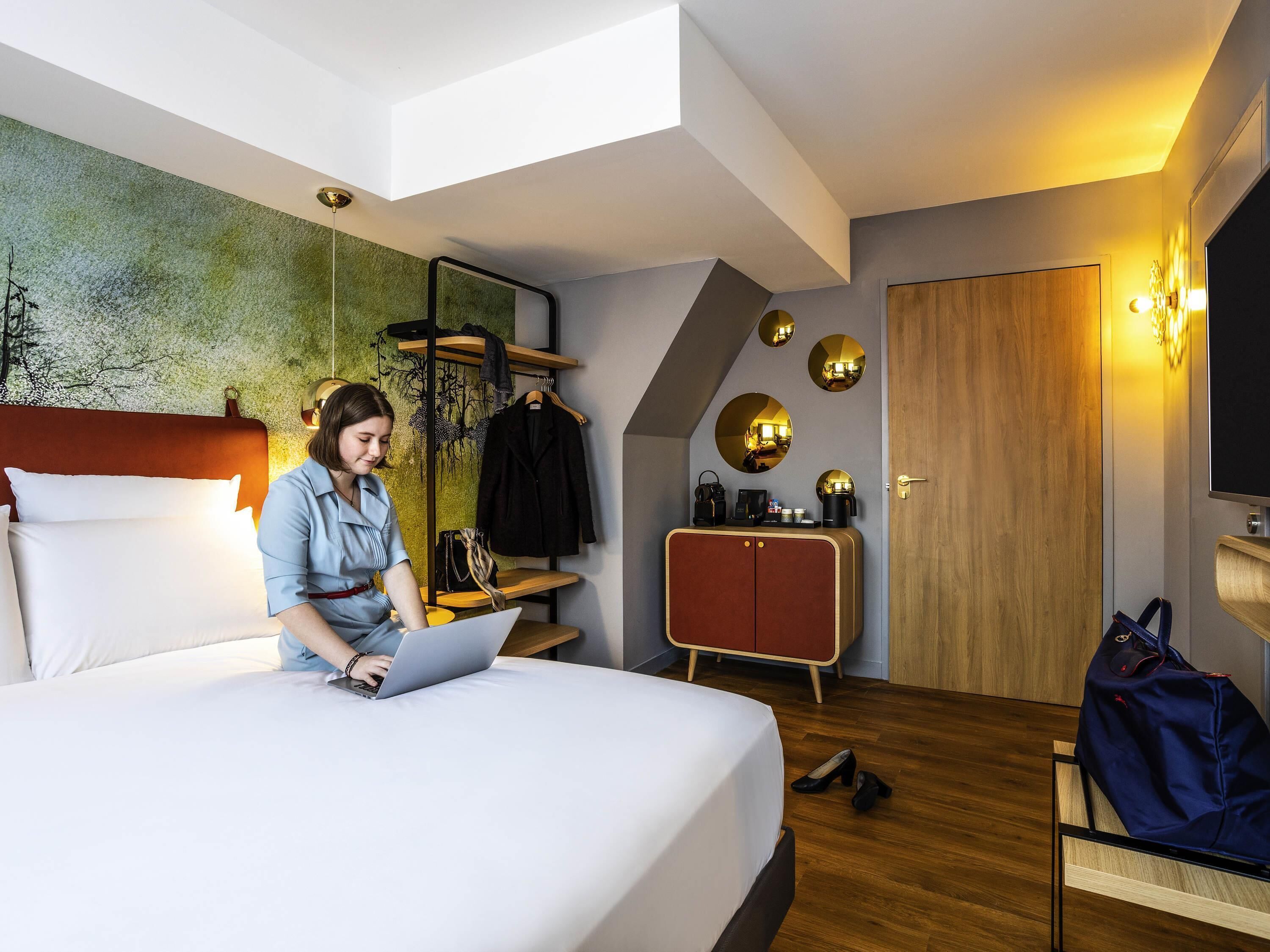 A modern hotel room with a bed, a laptop, a coat rack, a wardrobe, and a mirror.