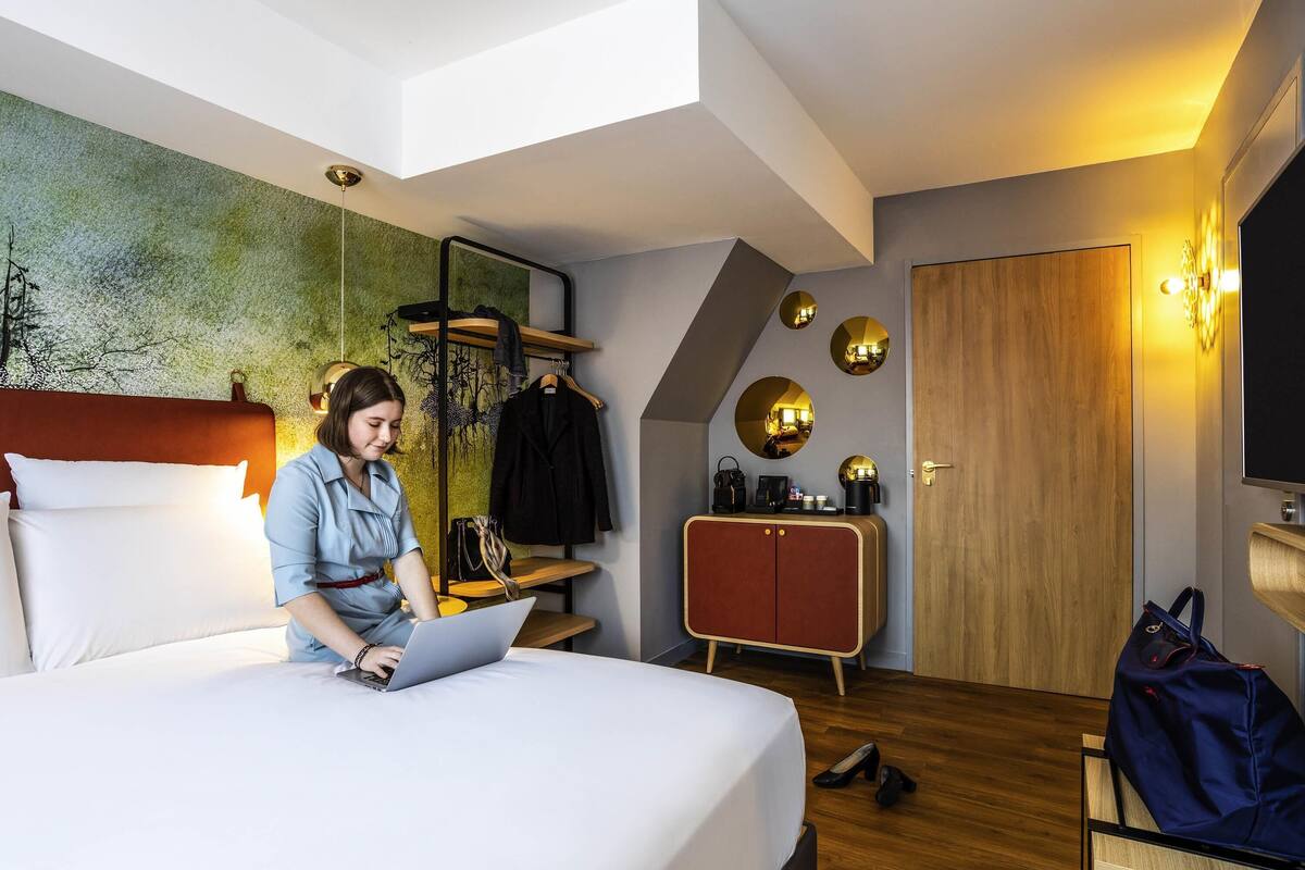 A modern hotel room with a bed, a laptop, a coat rack, a wardrobe, and a mirror.