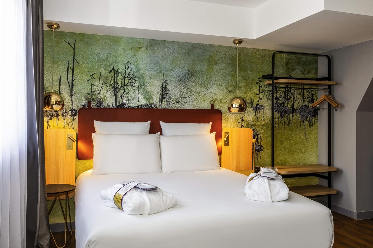 Habitación de hotel con una cama grande, cabecera, tapiz en la pared, mesita de noche, lámpara y perchero.